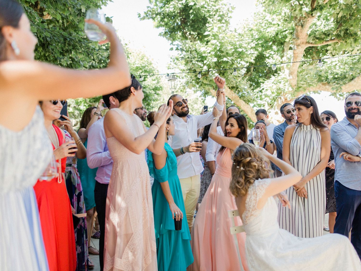 Guests dancing outdoors at a wedding celebration, wearing colorful formal attire, surrounded by trees.