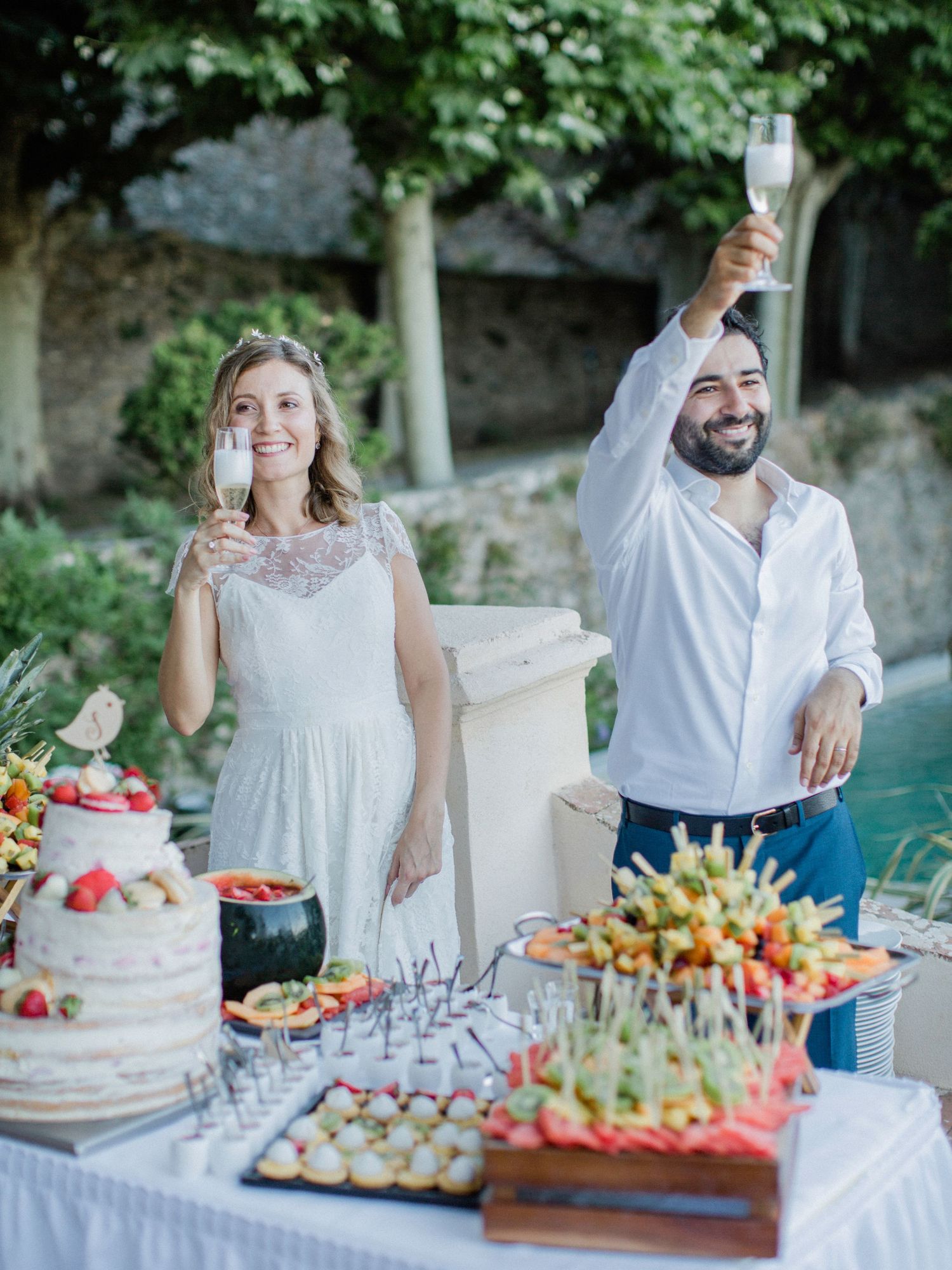 Bride and groom toasting with champagne glasses at elegant outdoor wedding reception with cake and fruit displays.