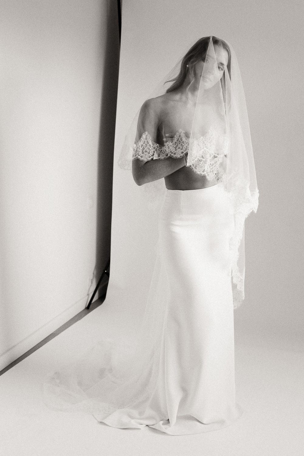 Black and white photo of a bride in an off-shoulder lace crop top, silk skirt, and flowing veil, posing elegantly.