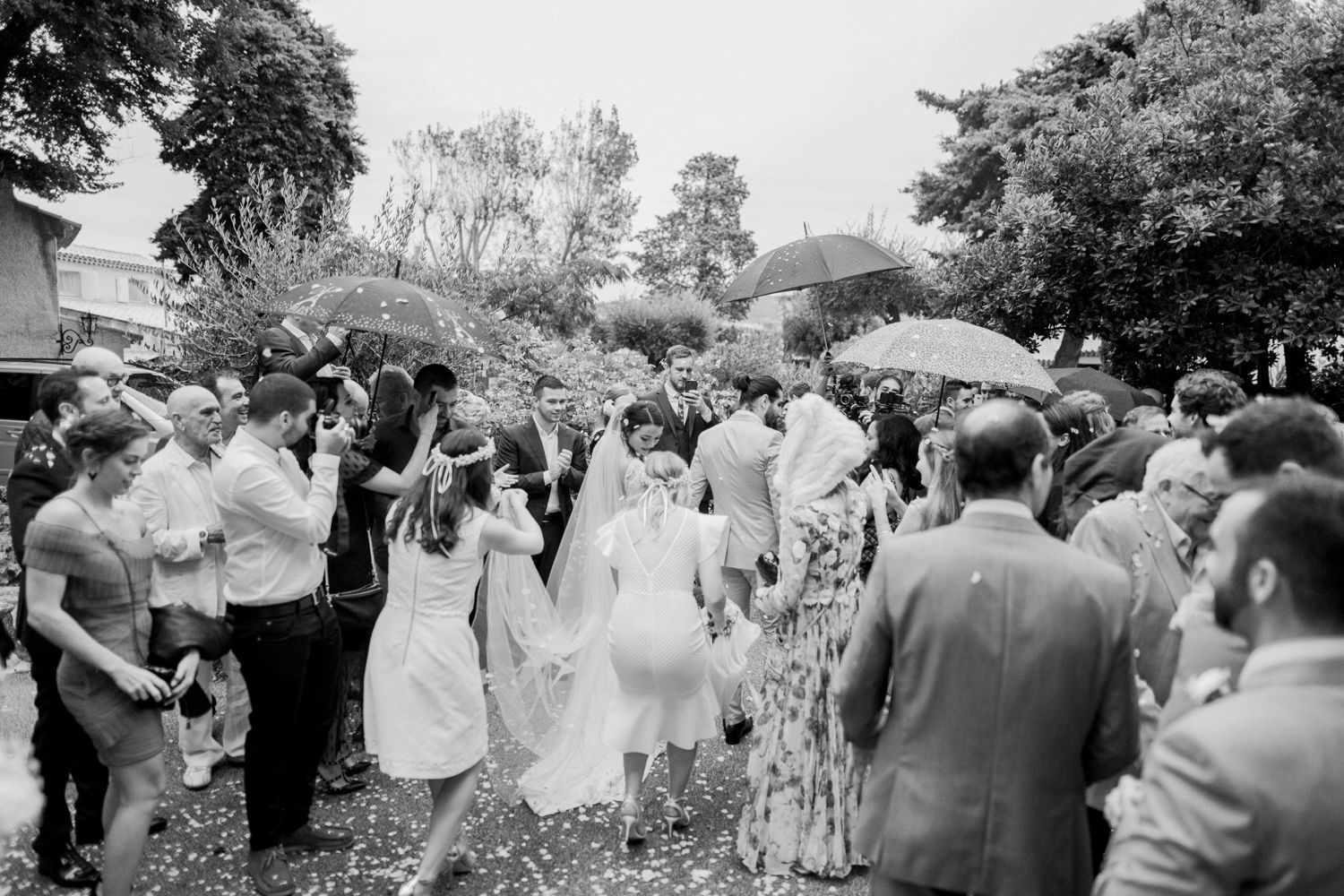 Black and white wedding photo of bride and groom walking through cheering guests outdoors.