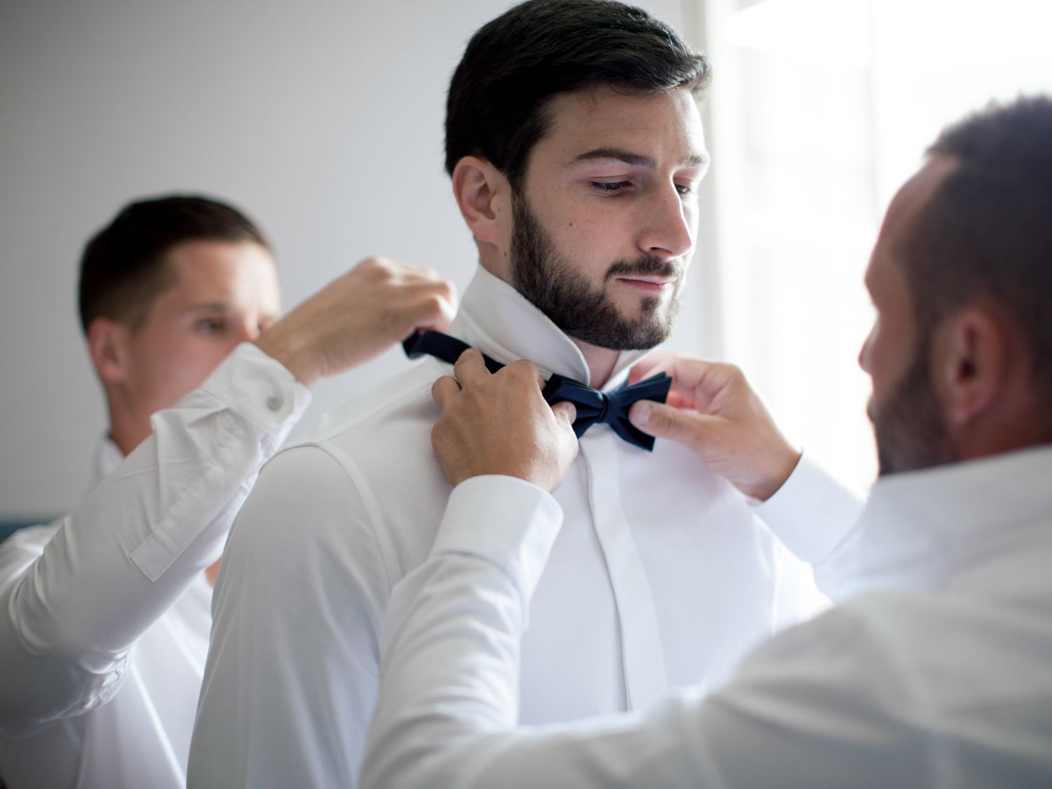 Two groomsmen helping groom adjust navy bow tie before wedding ceremony in white dress shirts.