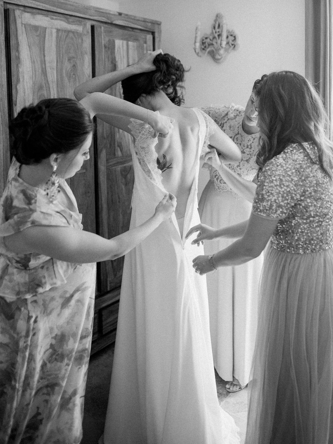 Black and white photo of bride being helped into wedding dress by two bridesmaids before ceremony.