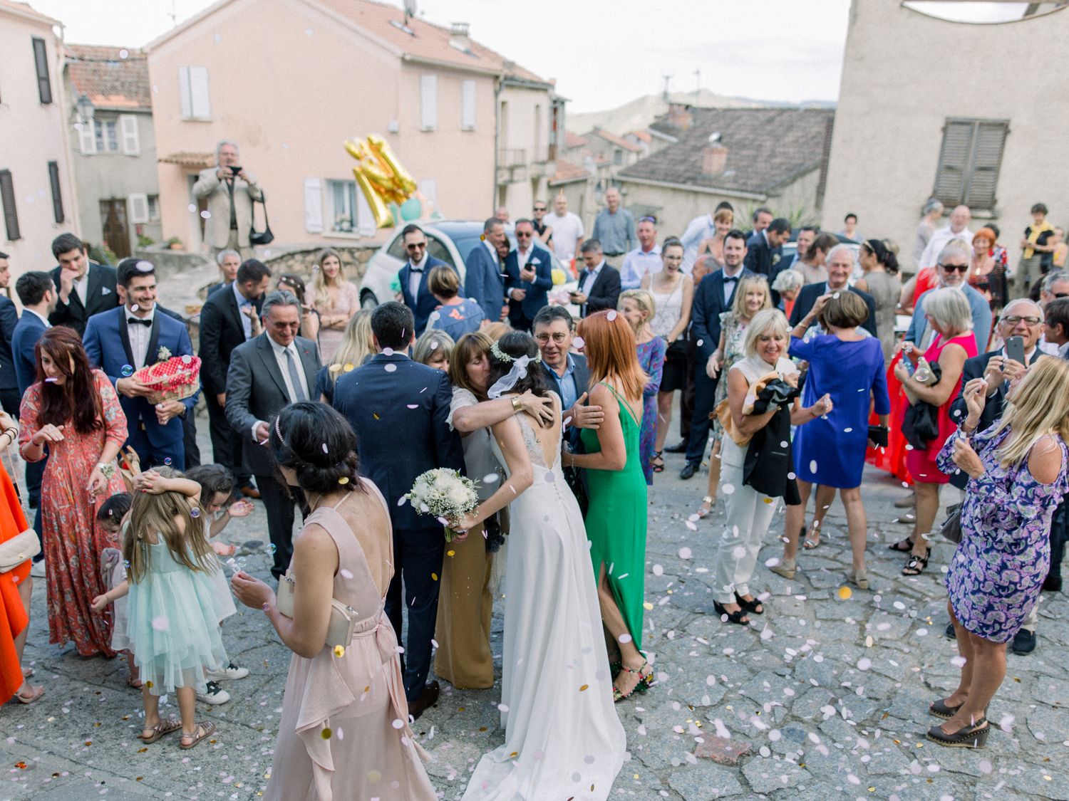 Bride and groom celebrate outdoors with guests throwing confetti at a joyful wedding reception in a charming European village square.