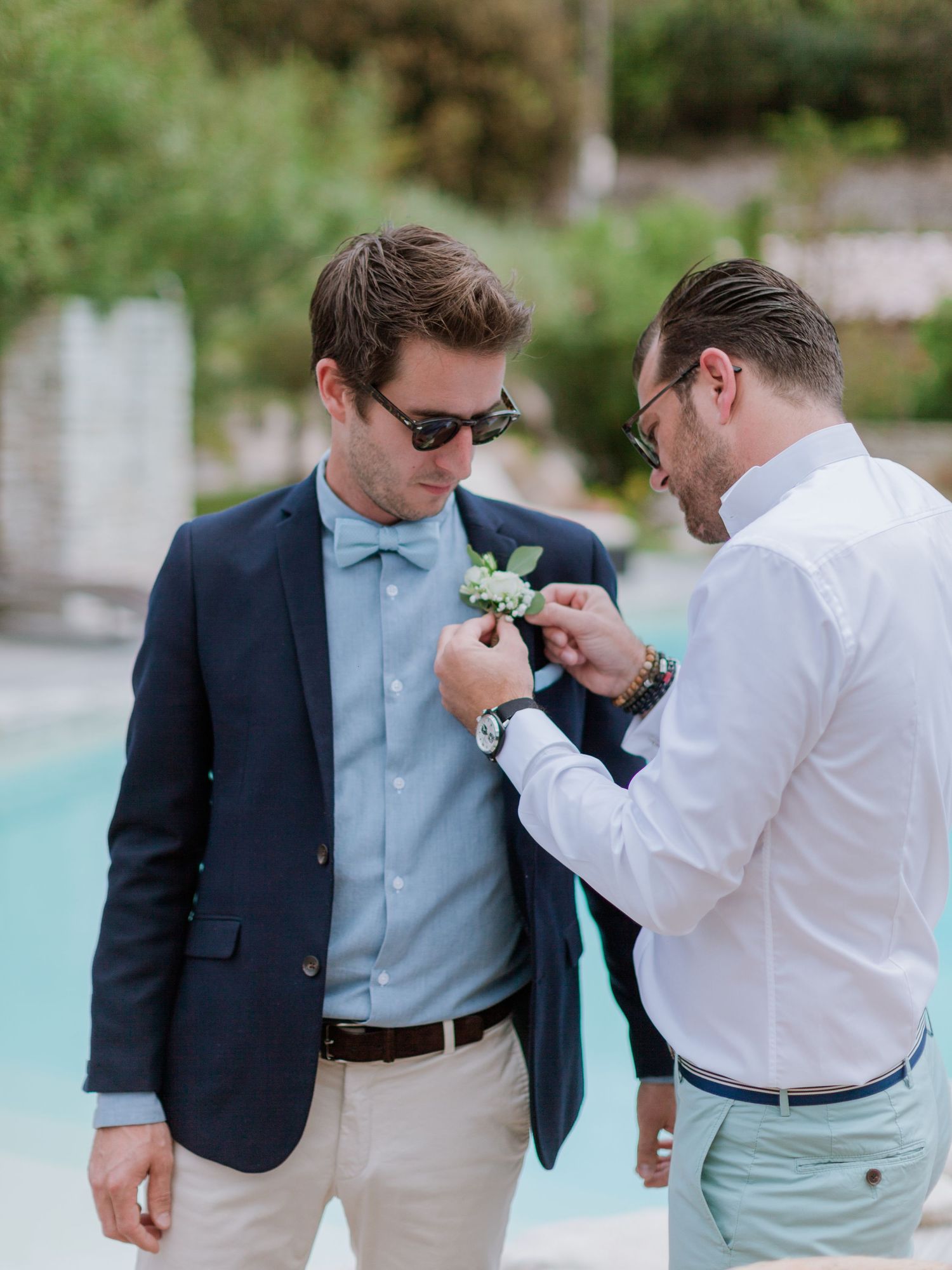 Un garçon d'honneur épingle une boutonnière au marié, vêtu d'un blazer bleu marine et d'un nœud papillon bleu, près d'une piscine.