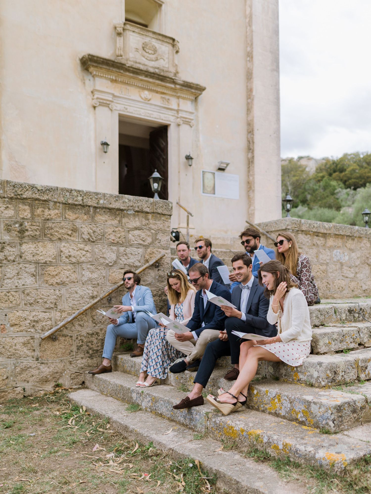 Des invités à un mariage, assis sur des marches en pierre devant une église à Bonifacio