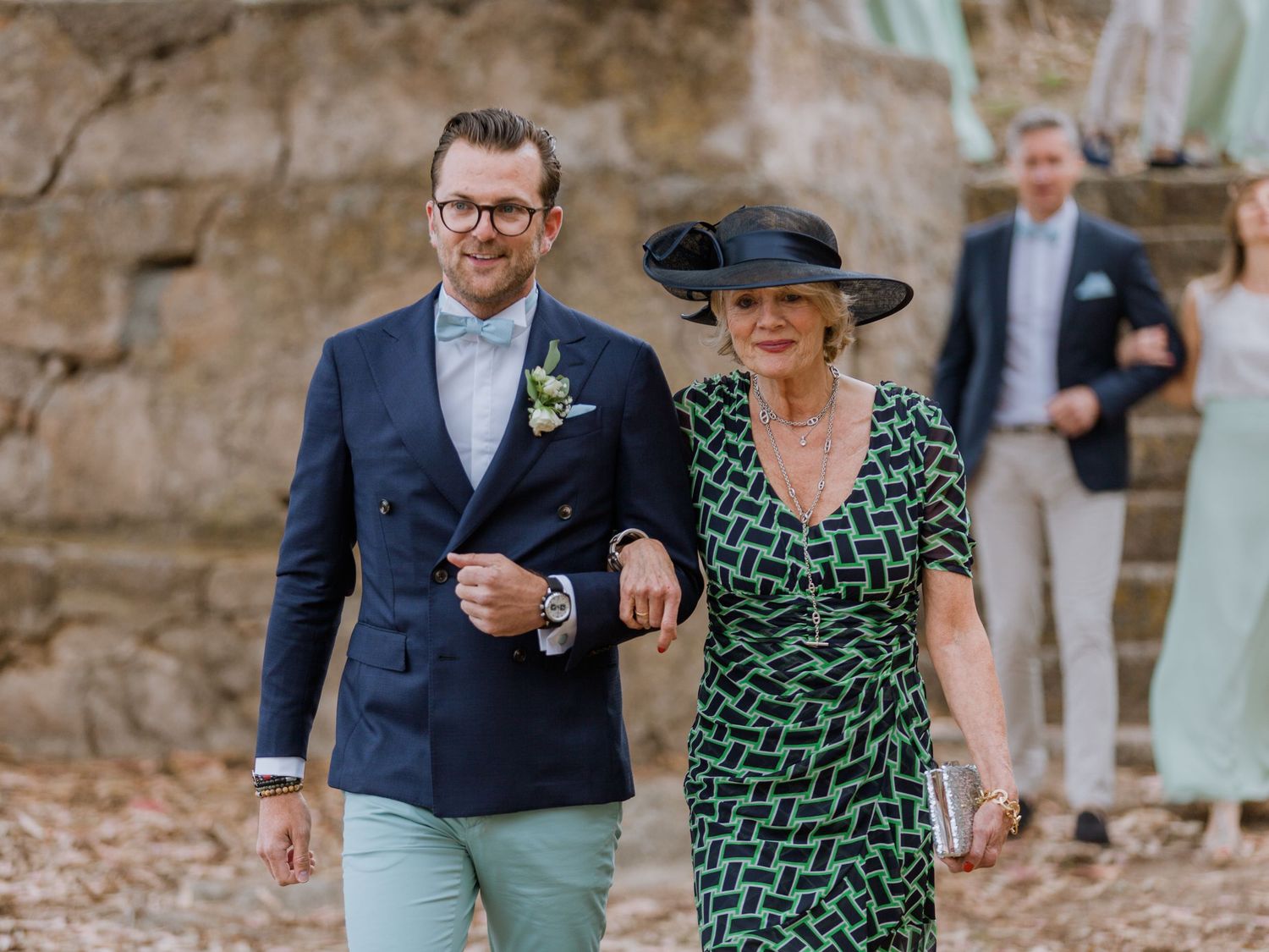 Un marié en costume bleu marine accompagne une élégante femme plus âgée en robe verte lors d'une cérémonie de mariage en plein air.
