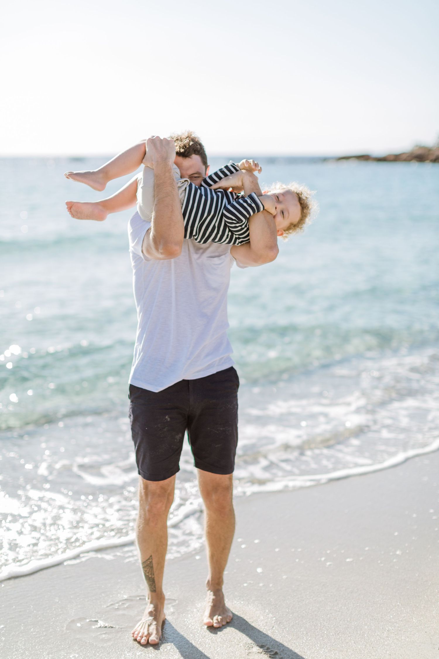 Un père soulève joyeusement son jeune enfant dans les airs sur une plage ensoleillée aux vagues turquoise de la mer à Palombaggia, Corse.