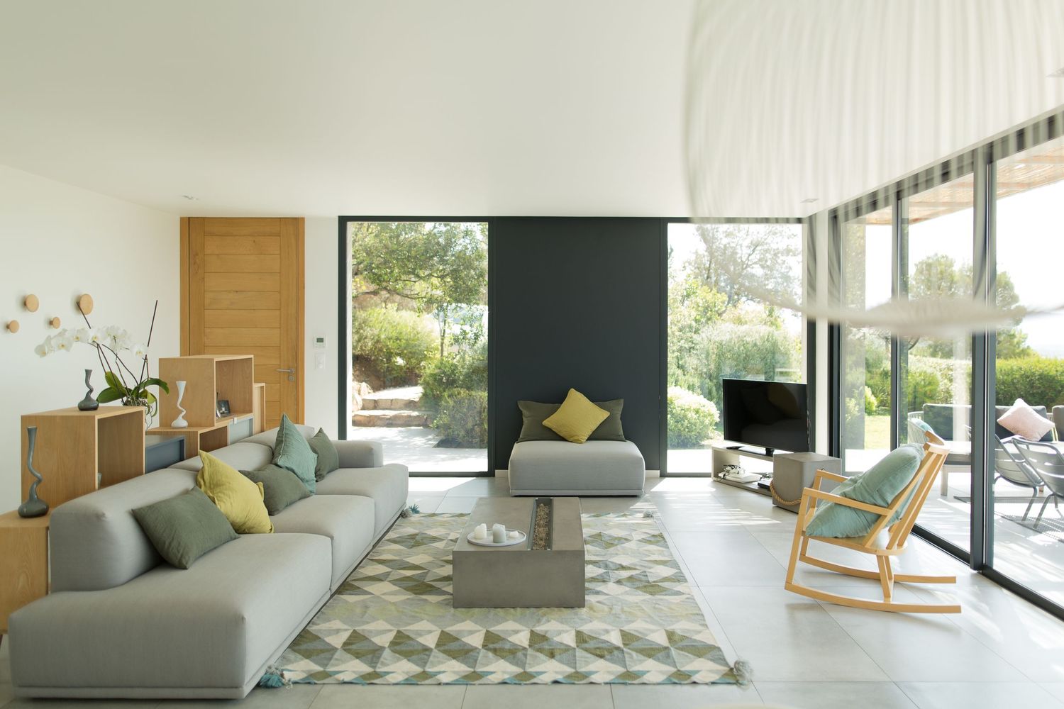 Salon lumineux et ouvert avec canapé d'angle gris, tapis géométrique, porte en bois et grandes portes vitrées donnant sur le jardin.