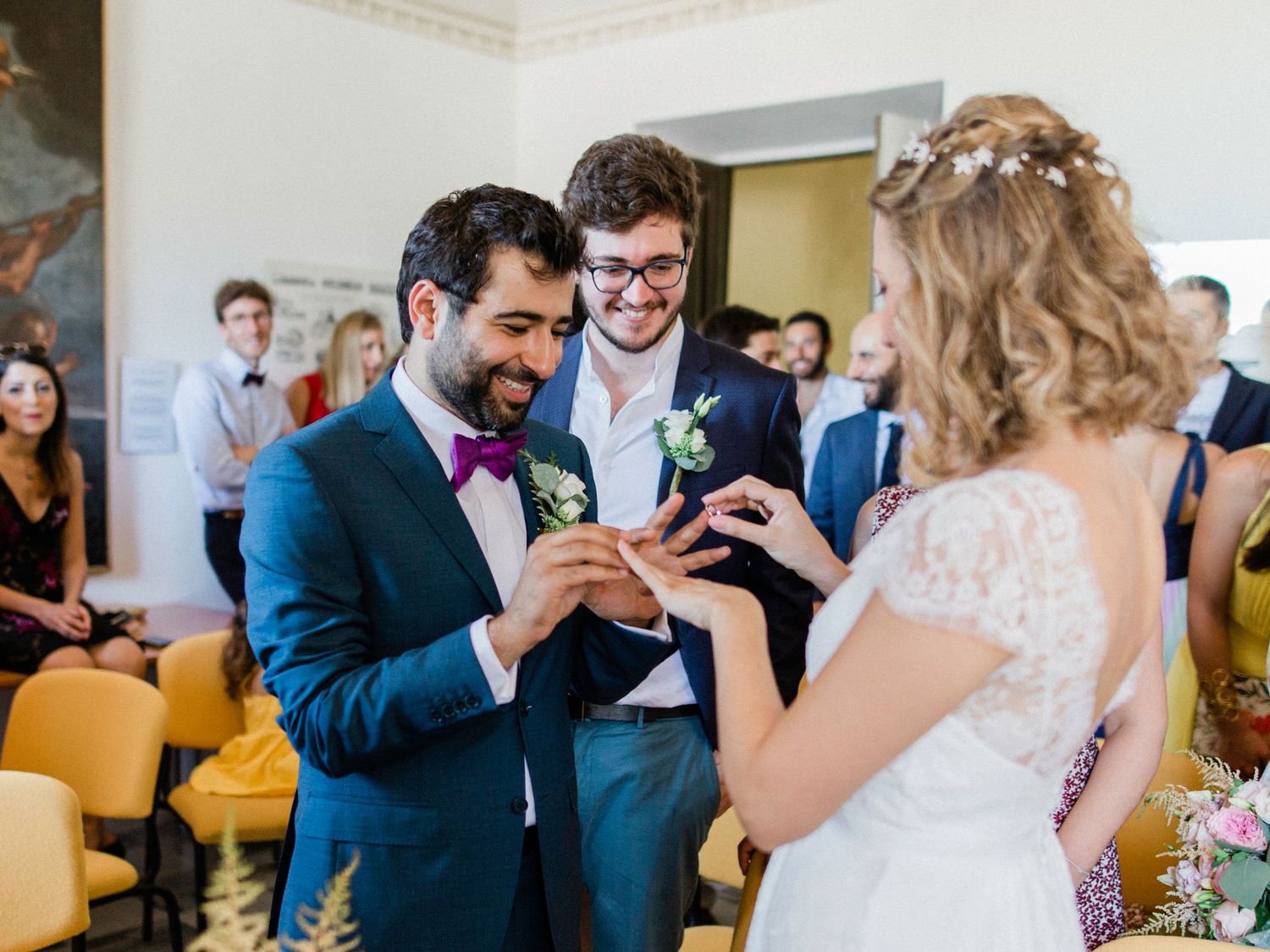 Groom places wedding ring on bride's finger during ceremony, officiant smiling, guests watching in elegant venue.