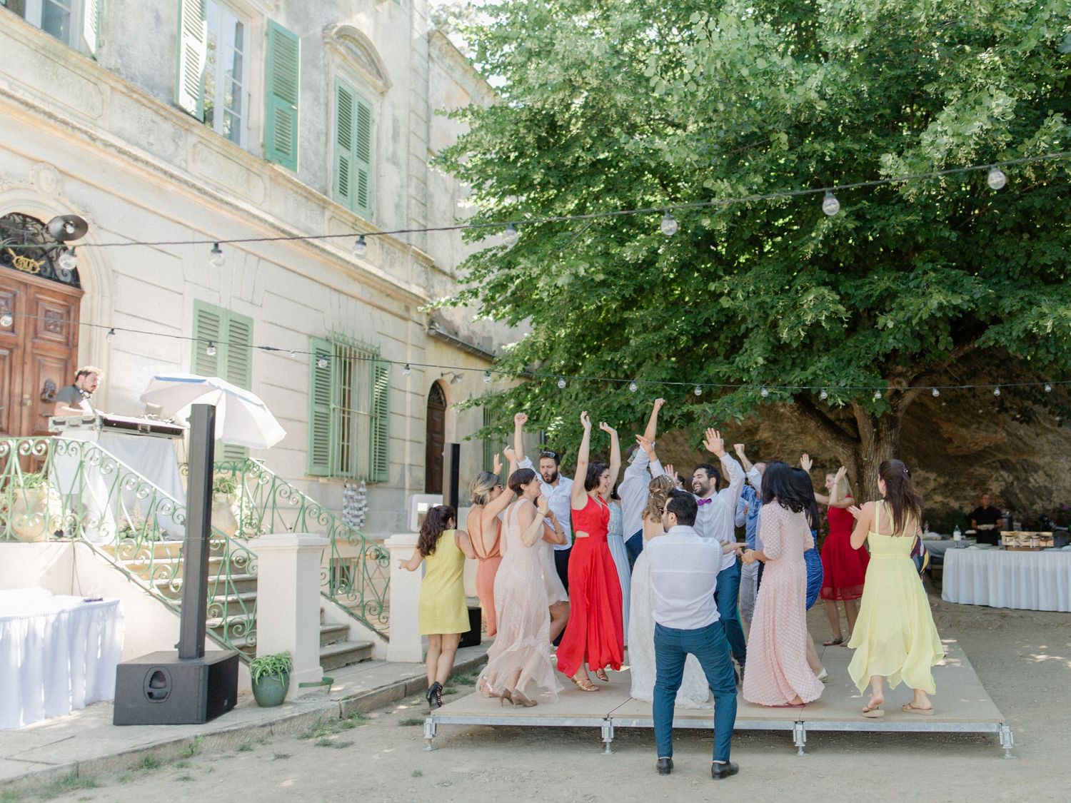 Wedding guests in colorful dresses dancing outdoors at elegant venue with lush green tree and historic building.