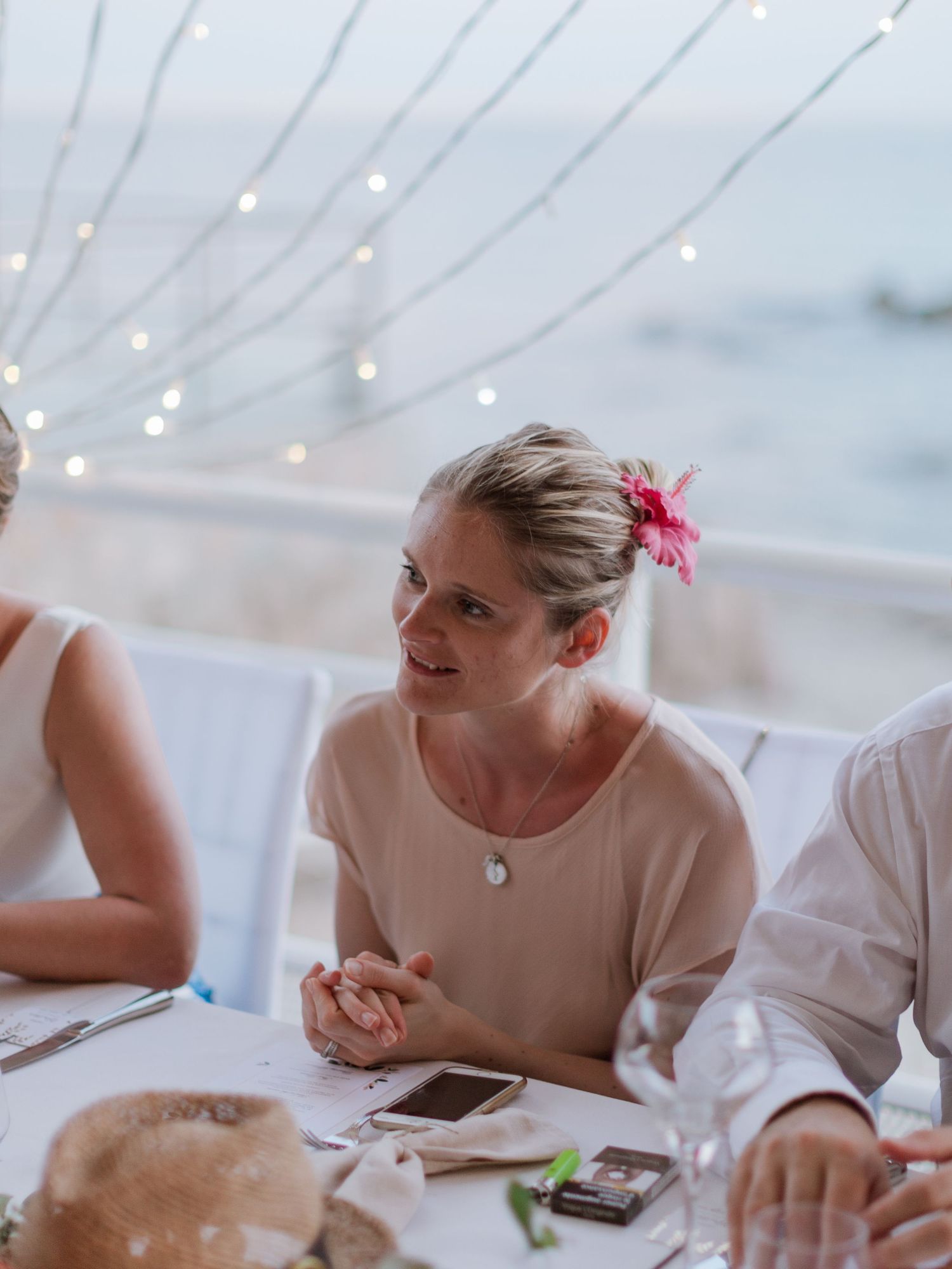 Une femme portant une fleur rose dans les cheveux est assise à une table de dîner en plein air, au bord de la plage, avec des guirlandes