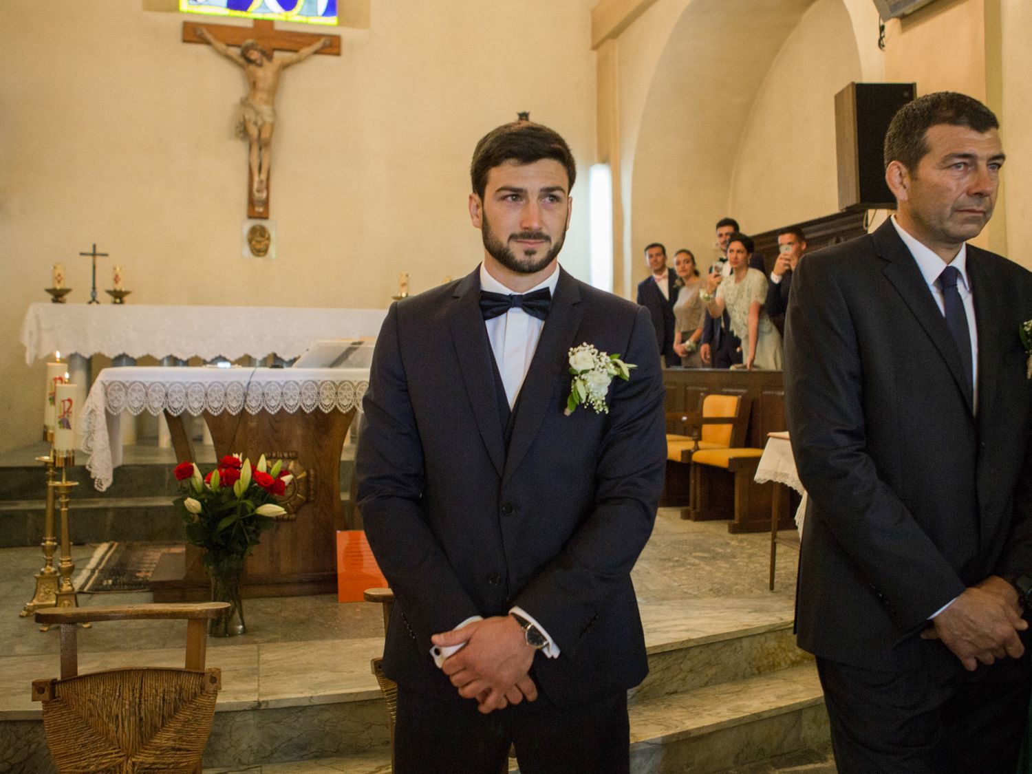 Groom in black tuxedo with boutonniere stands at church altar during wedding ceremony.