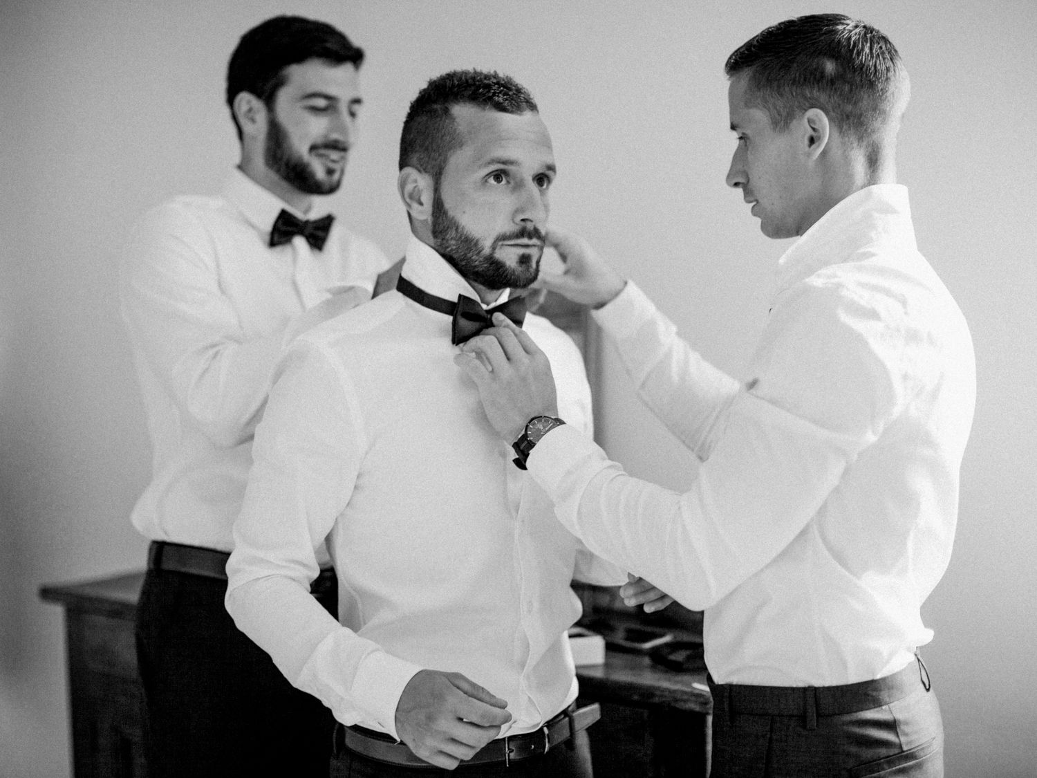 Black and white photo of groom getting bow tie adjusted by groomsmen before wedding ceremony.