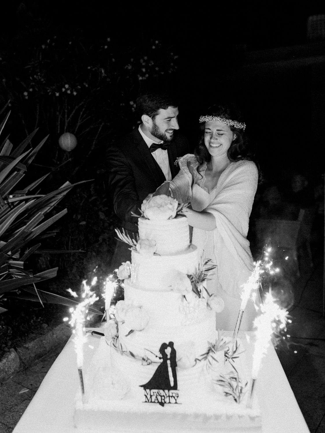 Les mariés coupent le gâteau de mariage avec des cierges magiques, photo en noir et blanc prise lors d'une réception en plein air en soirée.