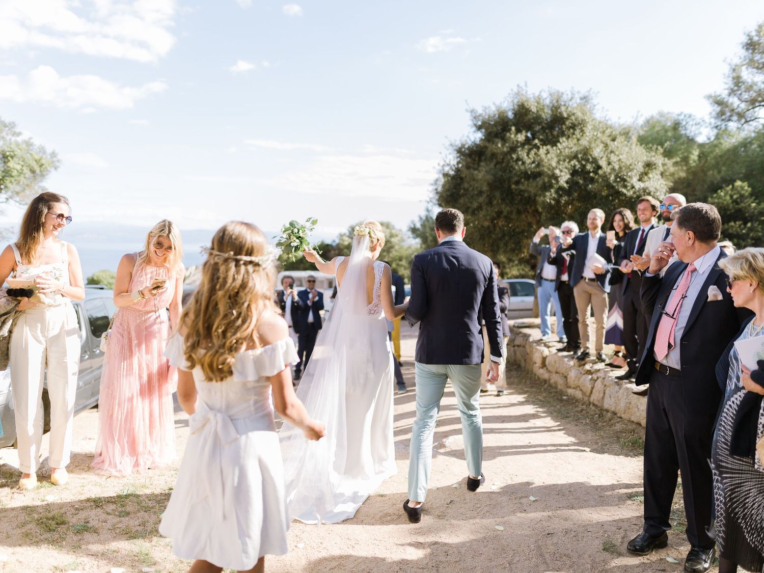 Les mariés traversent une foule d'invités enthousiastes lors d'une célébration de mariage ensoleillée, sous un ciel bleu et parmi les arbres