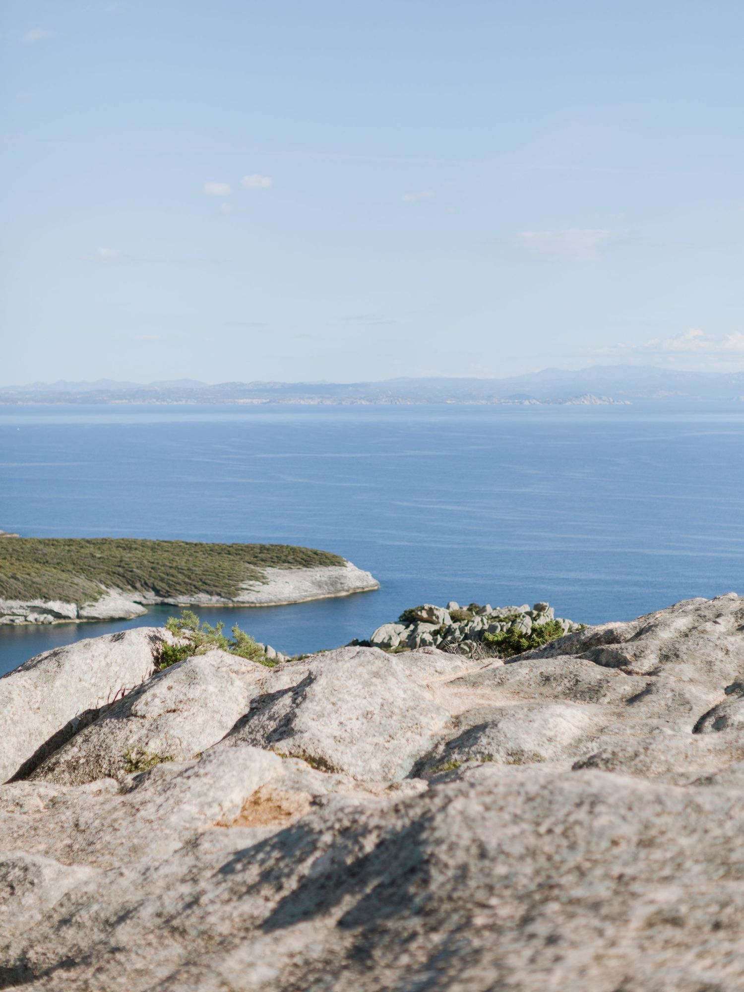 Côte rocheuse surplombant une mer bleue calme, avec une péninsule verdoyante et des montagnes au loin sous un ciel dégagé.
