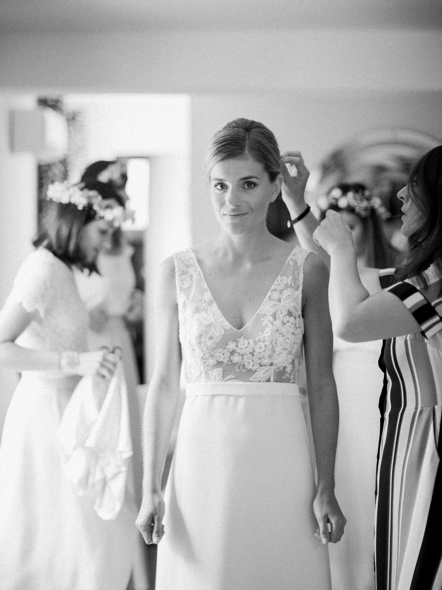 Photo en noir et blanc d'une mariée en robe de mariée en dentelle, aidée par ses demoiselles d'honneur coiffées de couronnes de fleurs.