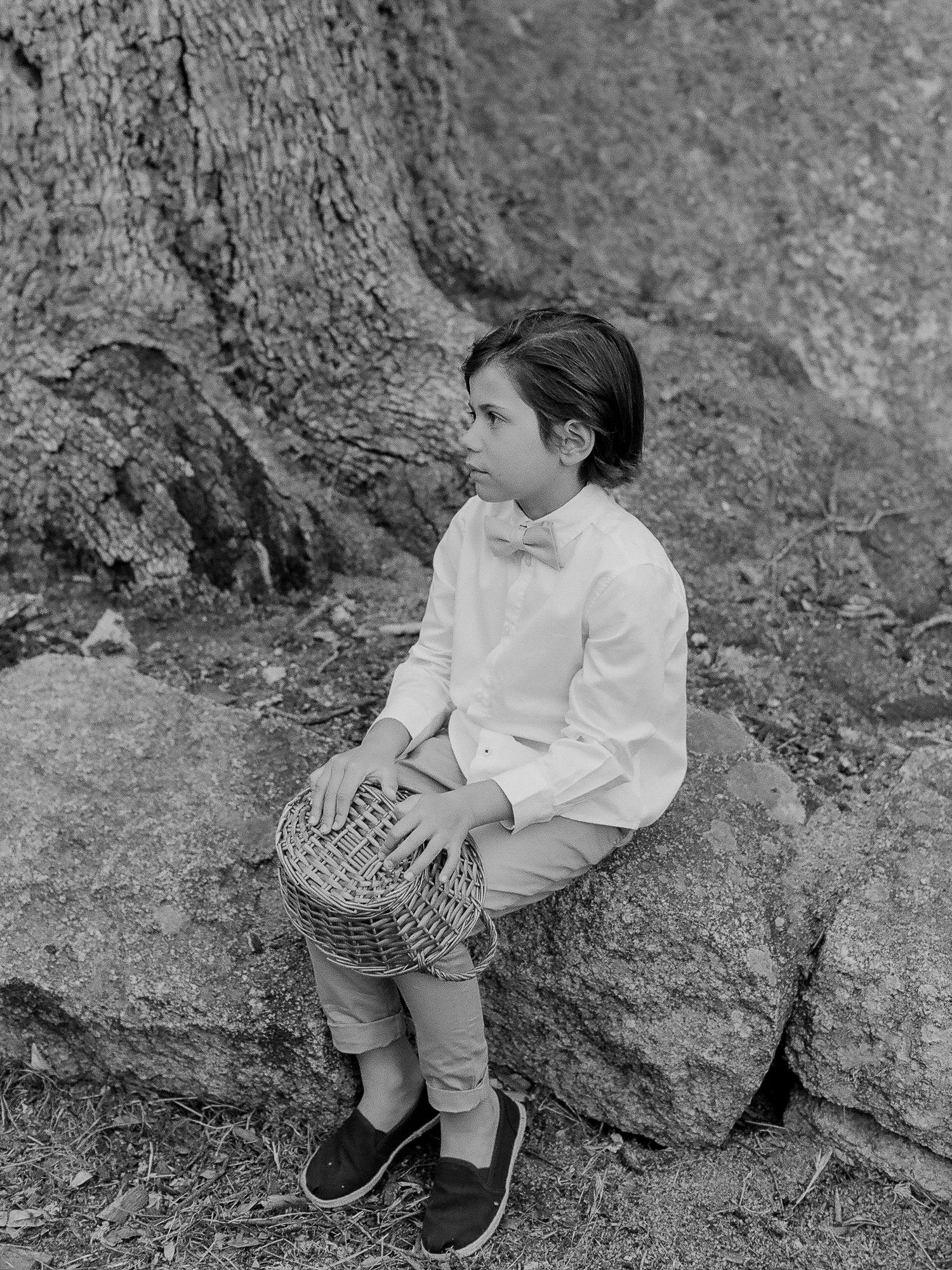 Un jeune garçon en chemise blanche et nœud papillon est assis sur un rocher, tenant un panier, posant près d'un arbre en noir et blanc.