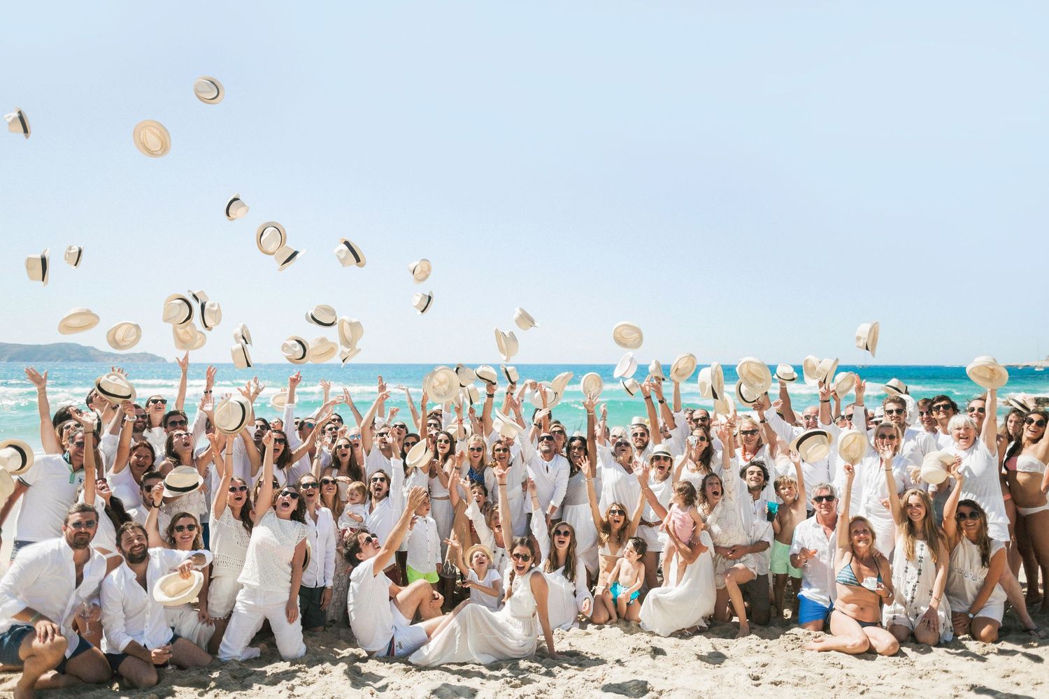 Grande photo de groupe de mariage sur la plage, tous les invités vêtus de blanc lançant des chapeaux de paille à calvi corse