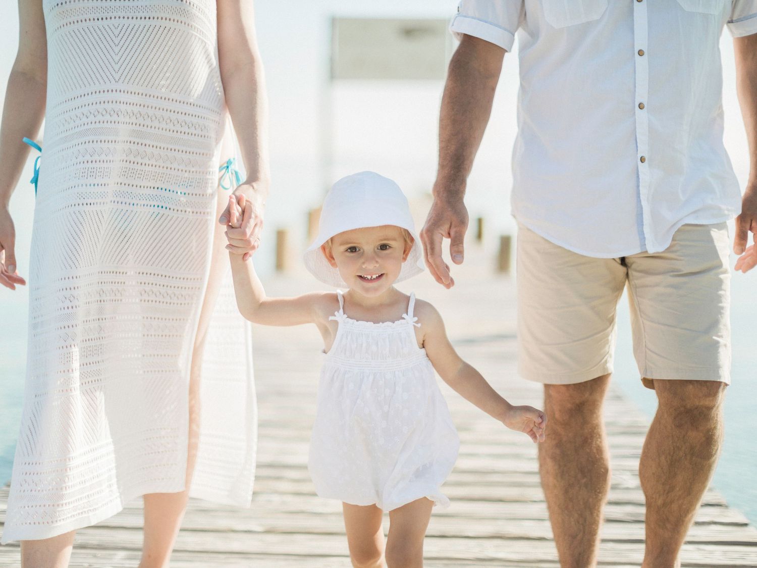 Un petit enfant souriant, vêtu d'une robe et d'un chapeau blancs, tient la main de ses parents en marchant sur une jetée ensoleillée.