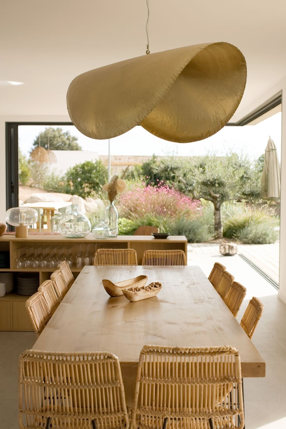 Salle à manger ouverte avec suspension sculpturale dorée, chaises en rotin et vue sur un jardin luxuriant à porto-vecchio