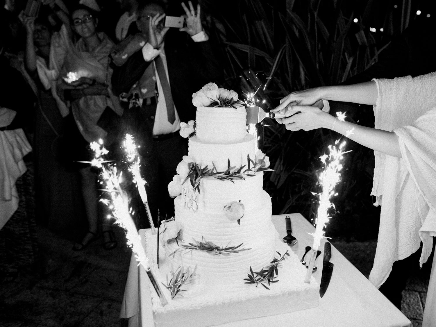 Photo en noir et blanc d'un gâteau de mariage à quatre étages entouré de cierges magiques, tandis que les mariés s'apprêtent à le couper.