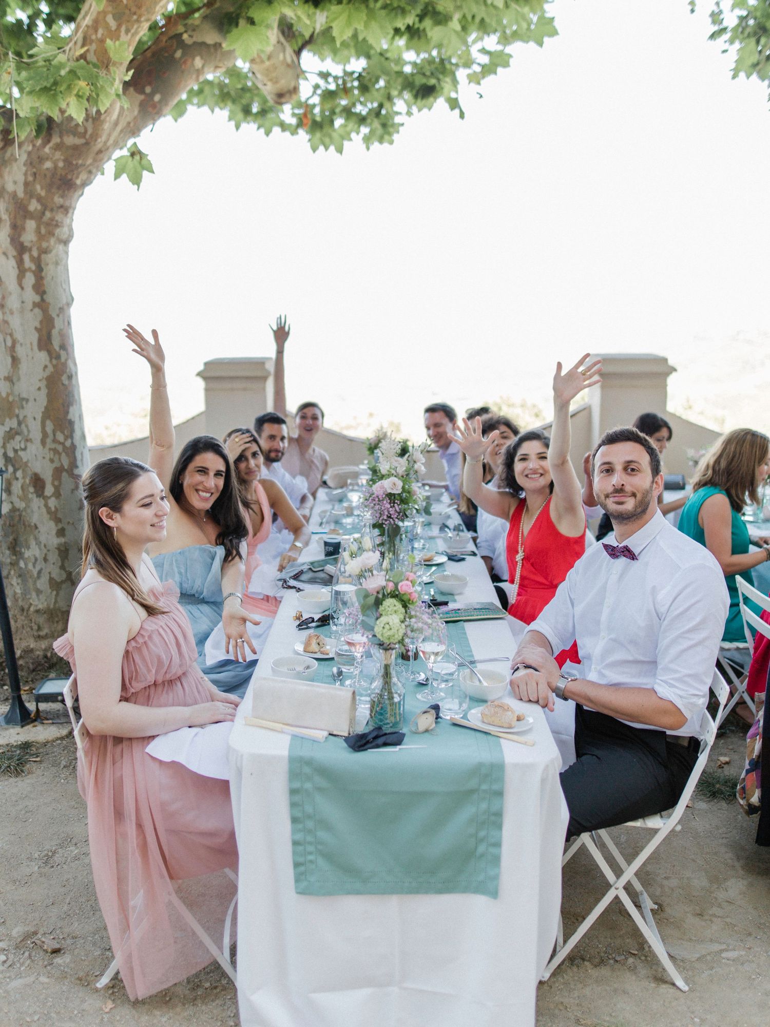Guests celebrate at an outdoor wedding reception dinner table under a tree, raising hands and smiling.