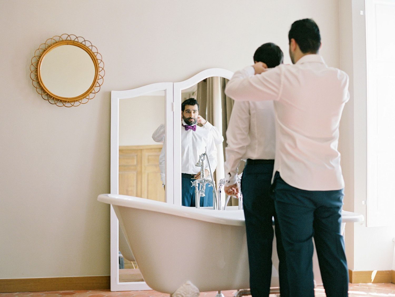 Two men stand before a white framed mirror, one helping the other with a tie in an elegant room.