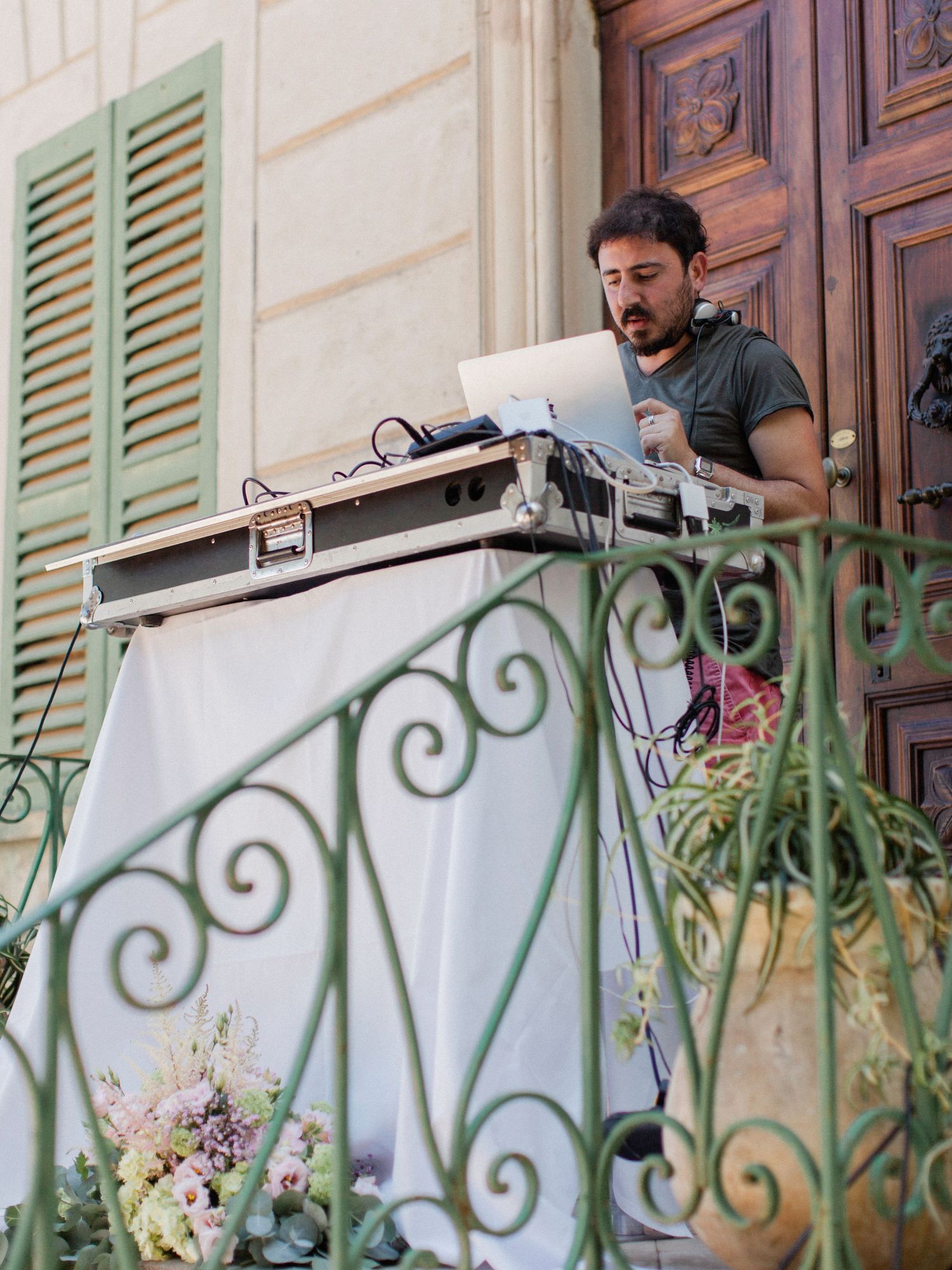 A DJ performs on an ornate balcony with a laptop and mixer, framed by green shutters and wrought iron railings.