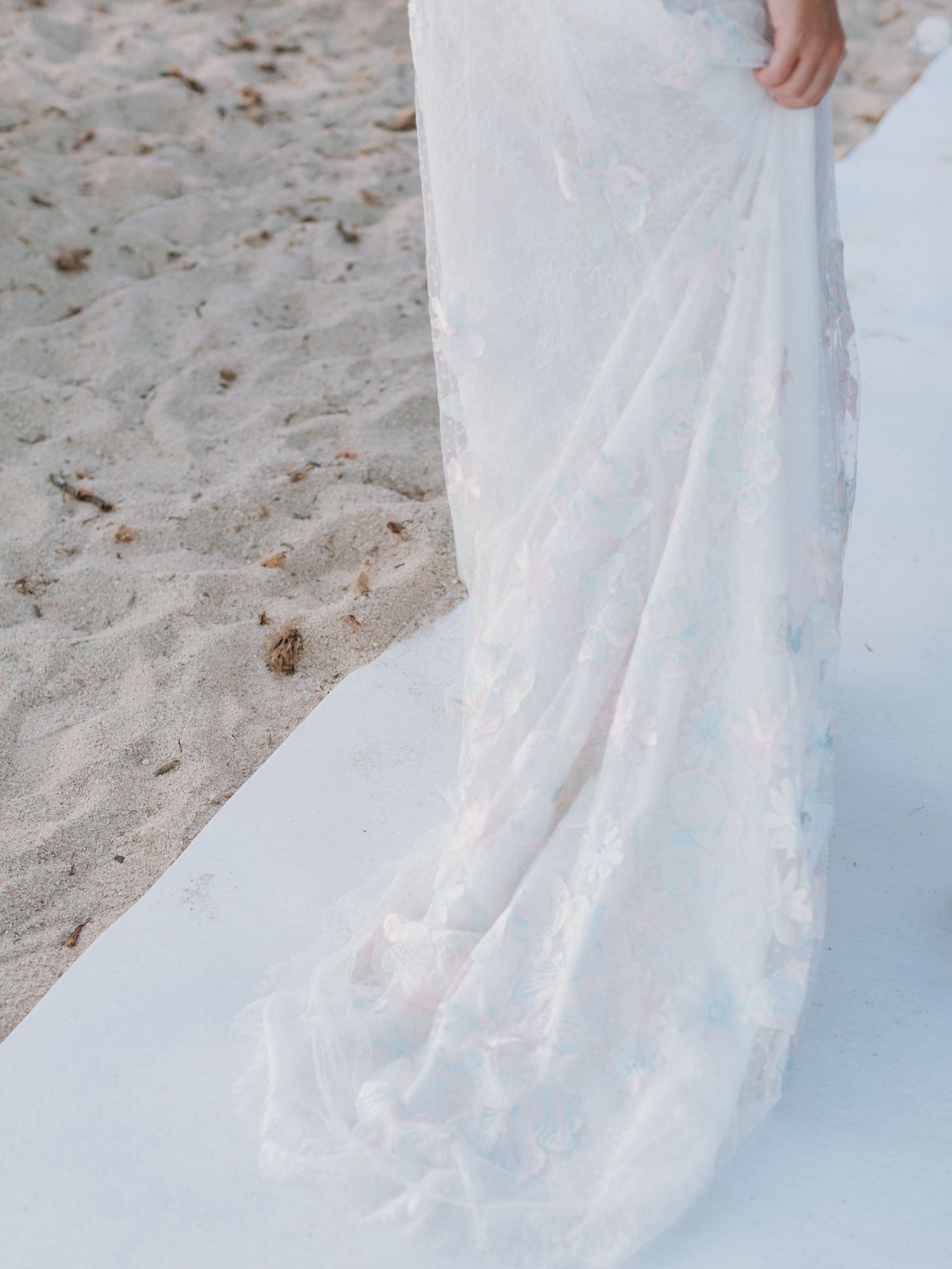 Traîne d'une élégante robe de mariée en dentelle florale étalée sur un tapis d'allée blanc, lors d'une cérémonie sur une plage de sable.