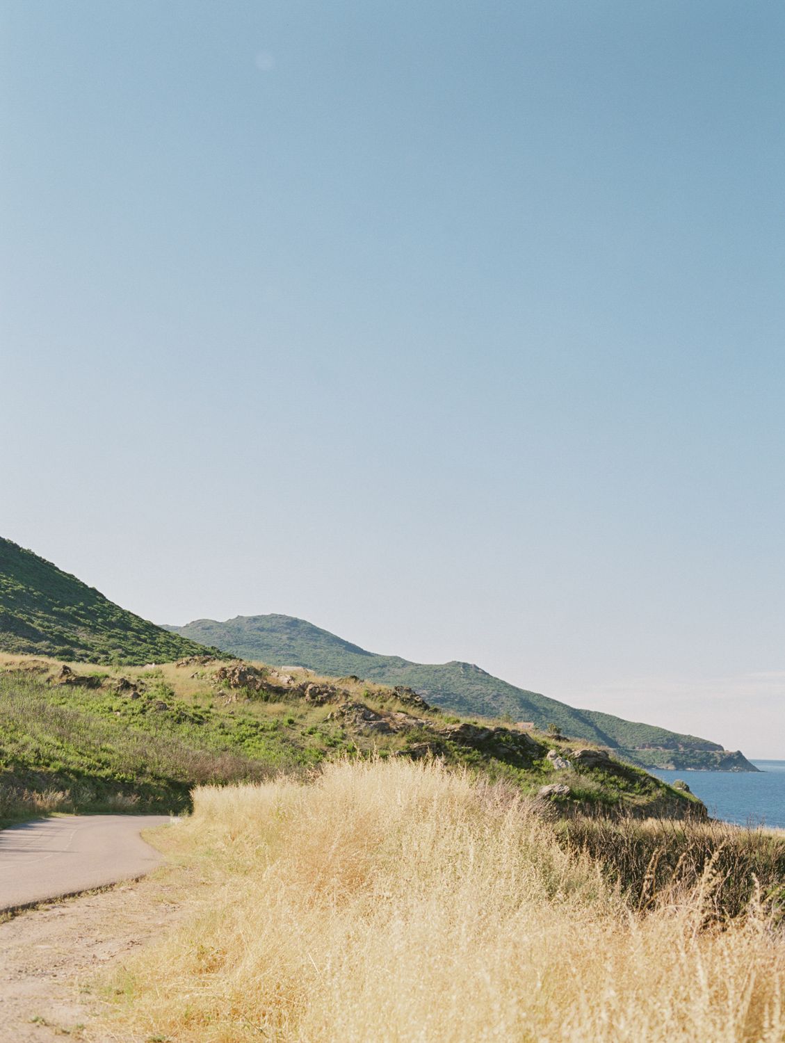 A scenic coastal path winds alongside golden dry grass and green hills overlooking a calm blue sea under a clear sky.