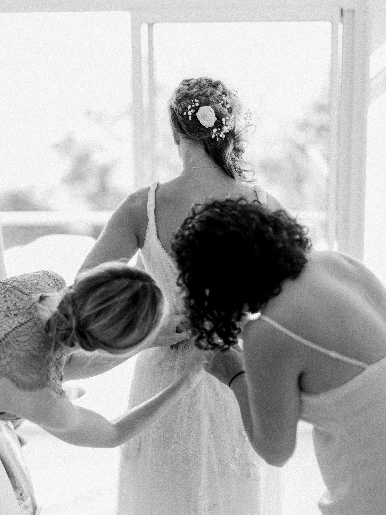 Photo en noir et blanc de demoiselles d'honneur aidant la mariée à enfiler sa robe près d'une fenêtre lumineuse avant le mariage.