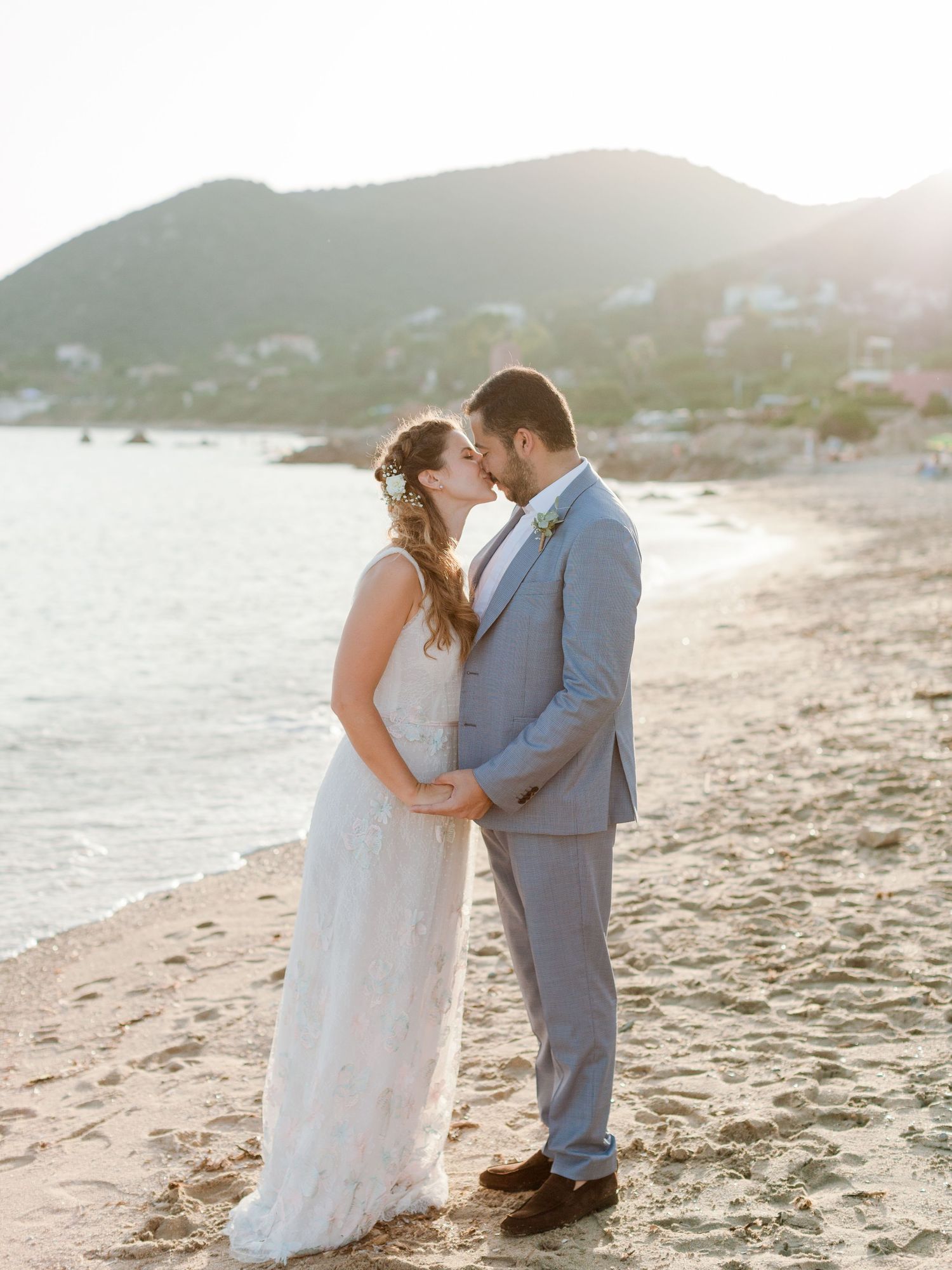 Un couple de jeunes mariés échange un baiser romantique sur une plage ensoleillée, avec les montagnes et l'océan en arrière-plan à Ajaccio