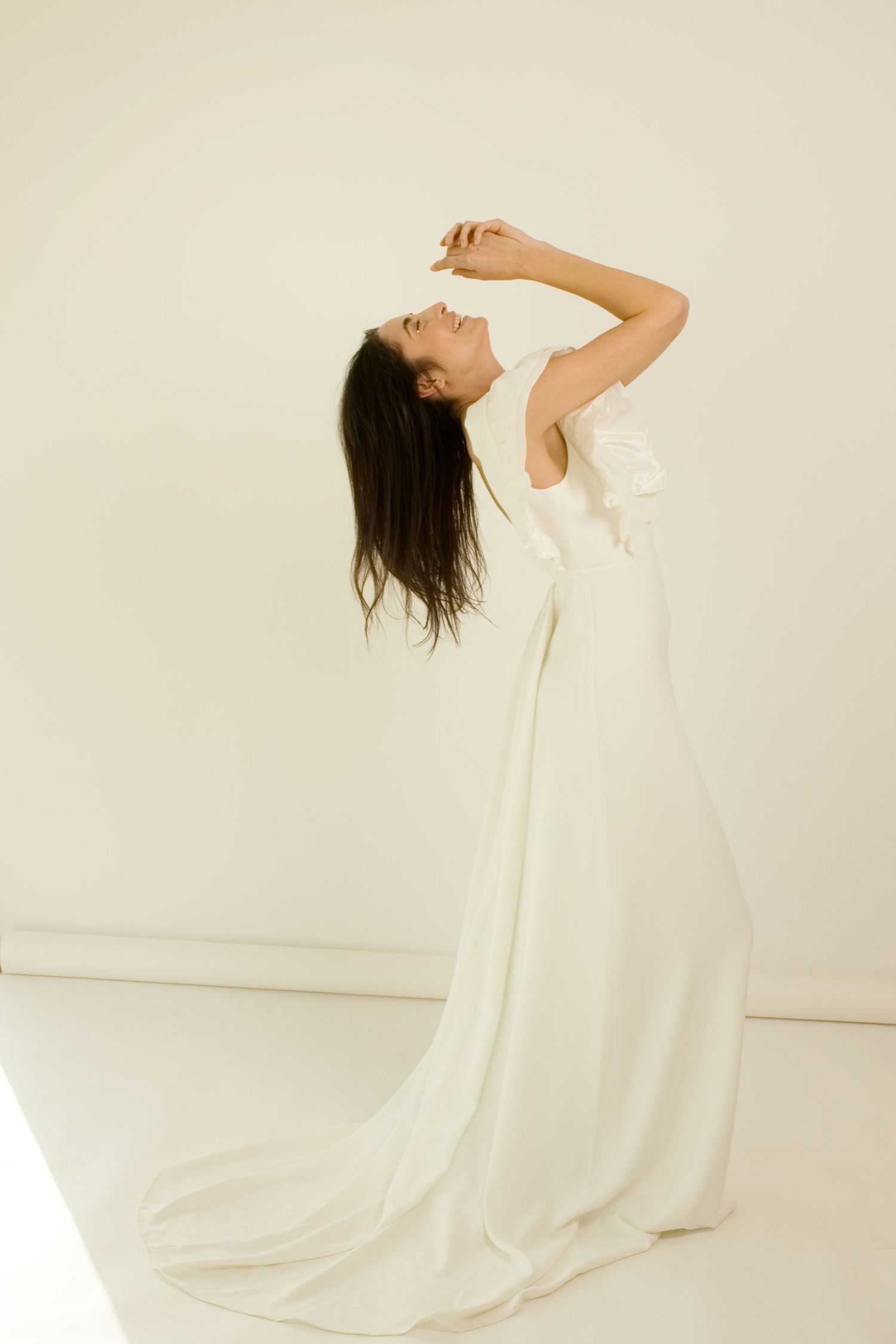 Femme en élégante robe de mariée blanche ornée d'un nœud, posant avec grâce, la tête renversée en arrière, dans un studio minimaliste.