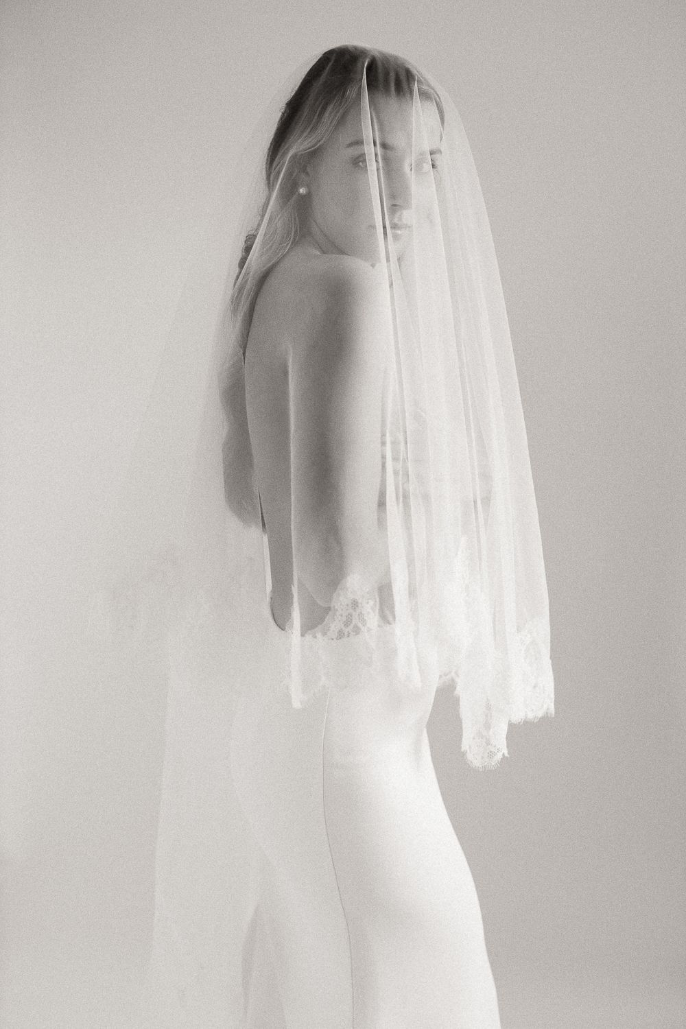 Black and white photo of a bride wearing a lace-trimmed veil and backless wedding gown, posed elegantly.