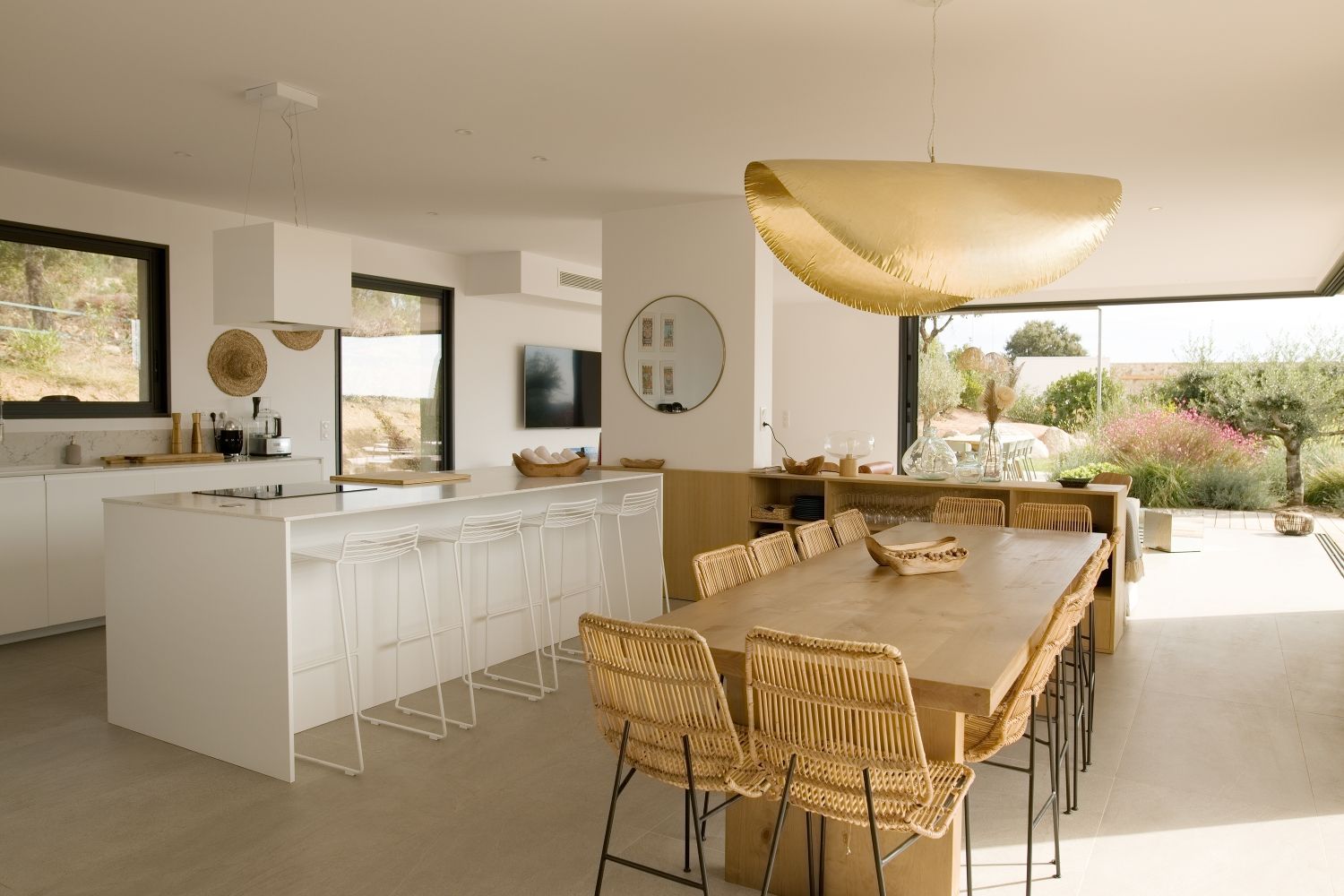 Cuisine et salle à manger lumineuses et ouvertes, avec îlot central blanc, chaises en rotin et suspension lumineuse dorée.