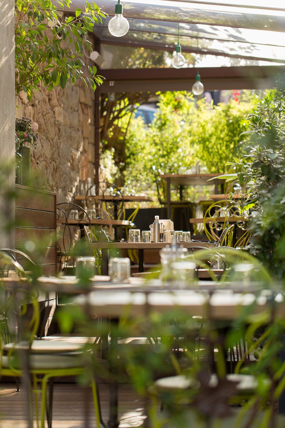 Terrasse ensoleillée d'un restaurant en plein air, agrémentée de plantes vertes, de guirlandes lumineuses et de tables en bois .