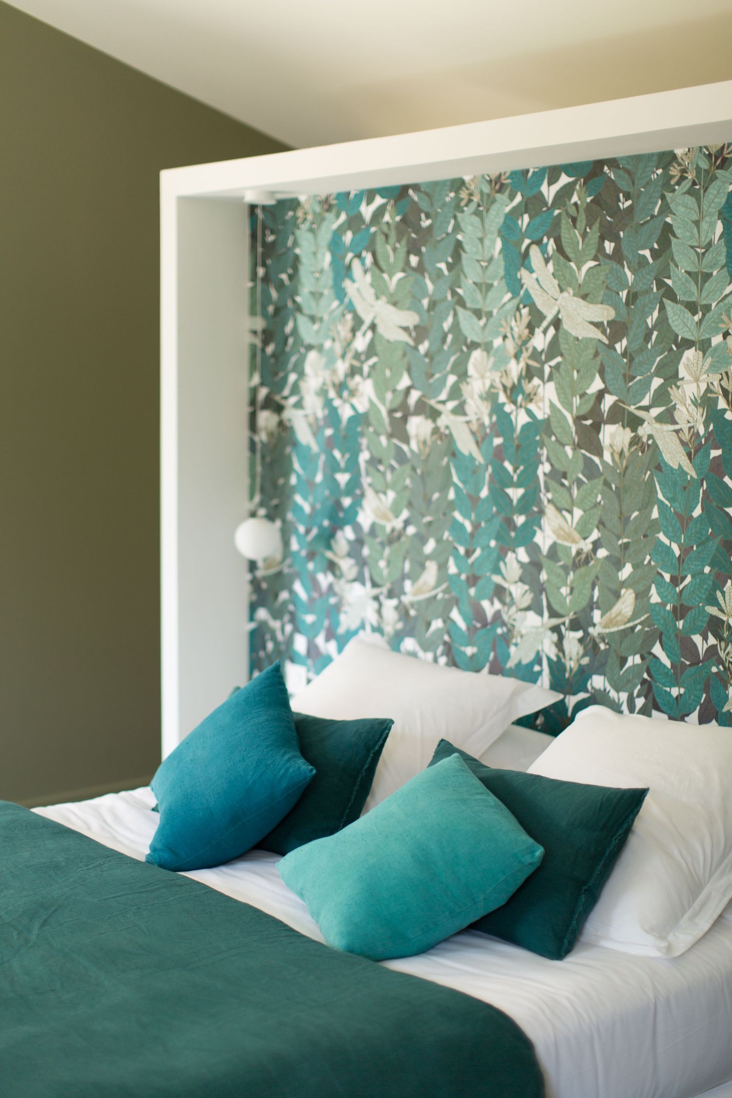 Chambre bleu sarcelle et blanche avec tête de lit ornée de papier peint à motifs floraux, coussins en velours turquoise et linge de lit.