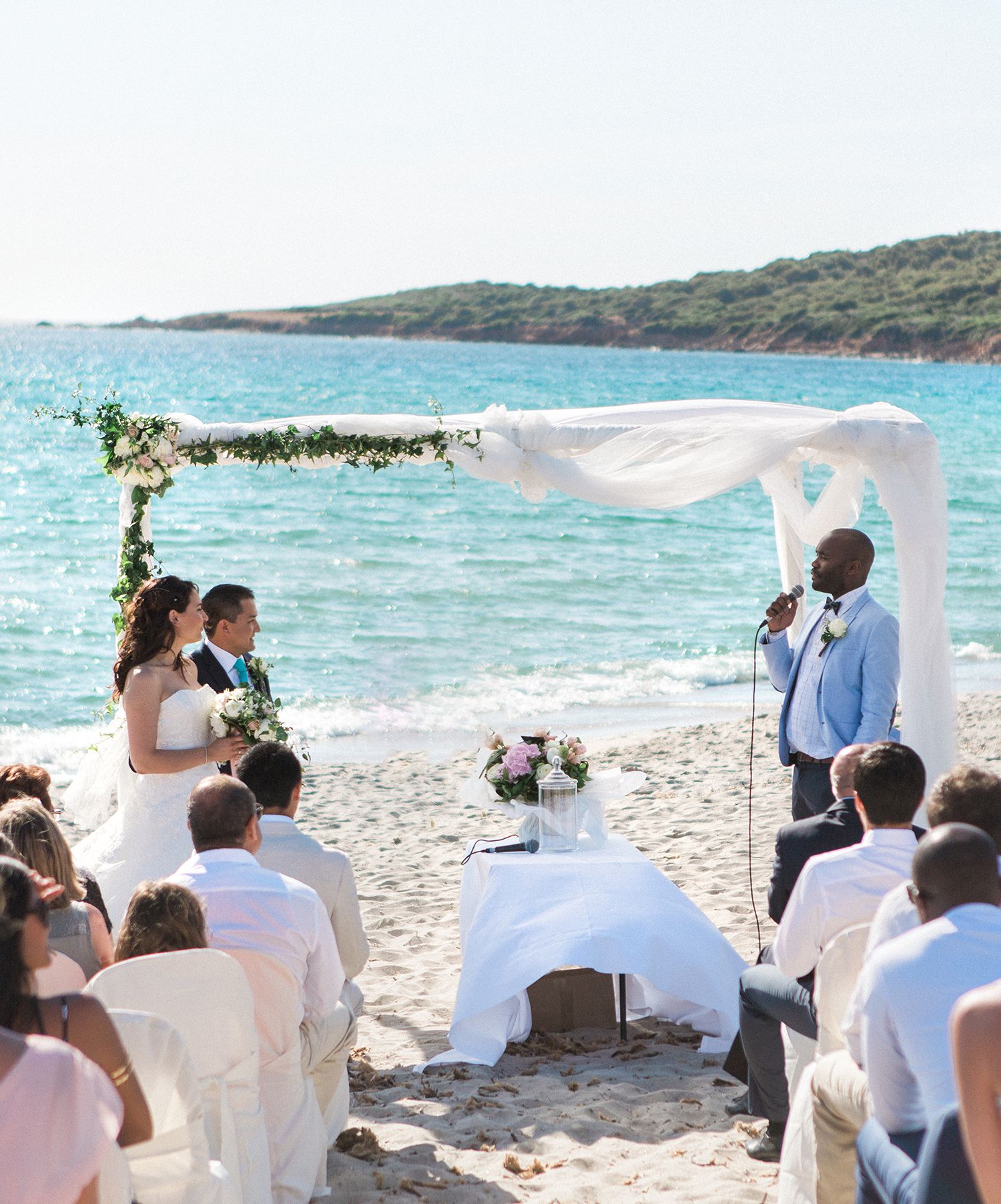 Cérémonie de mariage romantique sur la plage : les mariés sous une arche fleurie, l'officiant en costume bleu