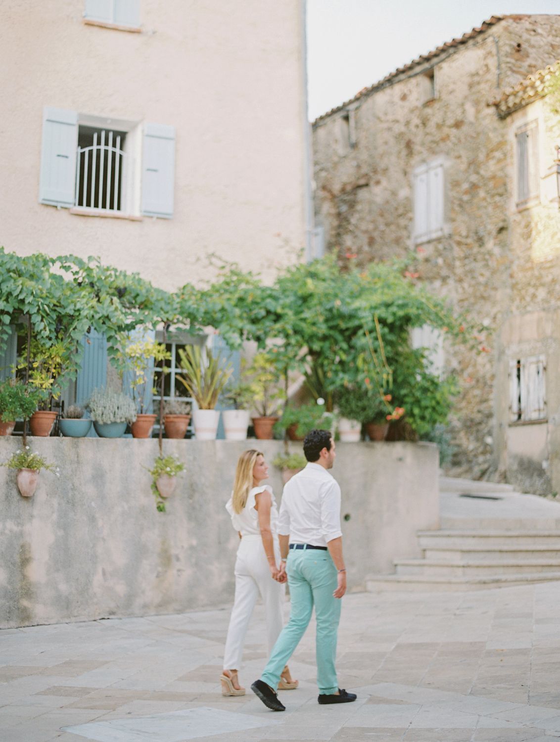 Un couple se tenant la main traverse la charmante cour en Provence, bordée de bâtiments en pierre et de verdure luxuriante.