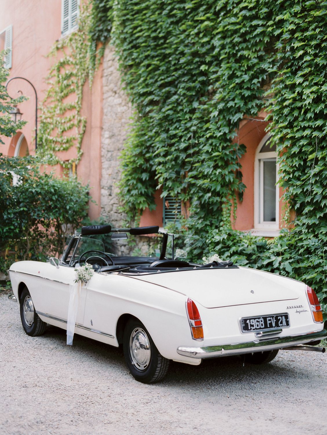 Vintage 1968 white convertible wedding car decorated with flowers, parked outside ivy-covered pink building in France.