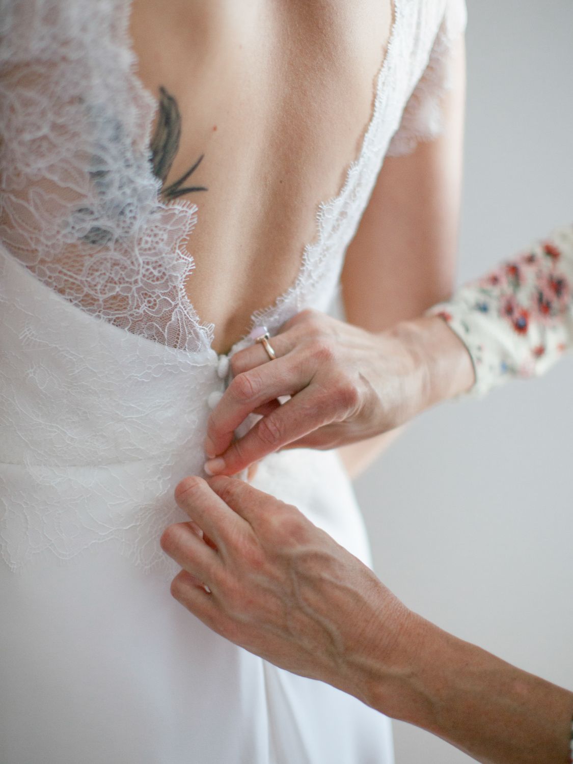 Close-up of hands fastening buttons on a white lace wedding dress with a low back and tattoo visible.