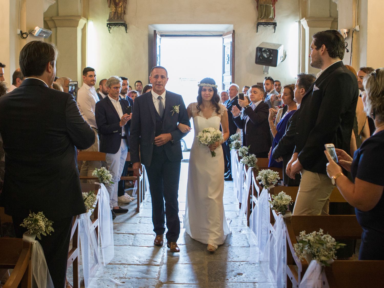 Bride and groom walk down church aisle surrounded by smiling guests at a sunlit wedding ceremony.