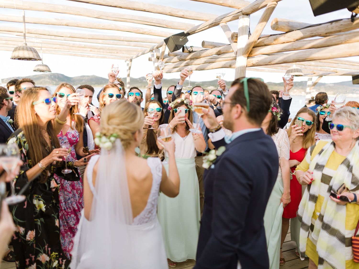 Les jeunes mariés sont célébrés par leurs invités, lunettes de soleil sur le nez, levant leurs verres à l'extérieur sous une pergola