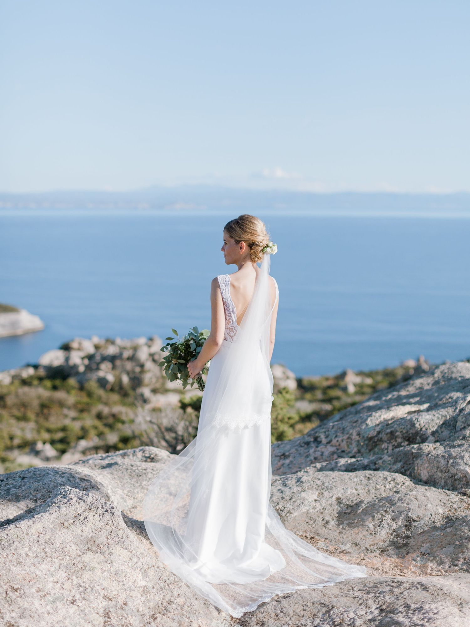 Une mariée en robe blanche, tenant son bouquet, se tient sur une falaise rocheuse surplombant la mer à Bonifacio en corse