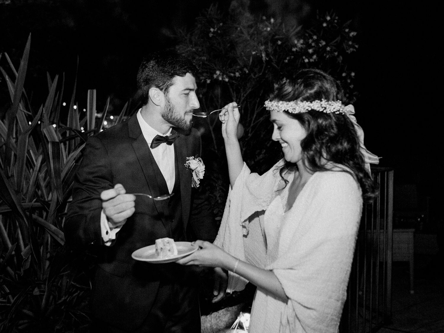 Photo en noir et blanc d'une mariée donnant à manger au marié le gâteau de mariage lors d'une réception de mariage en plein air, de nuit.