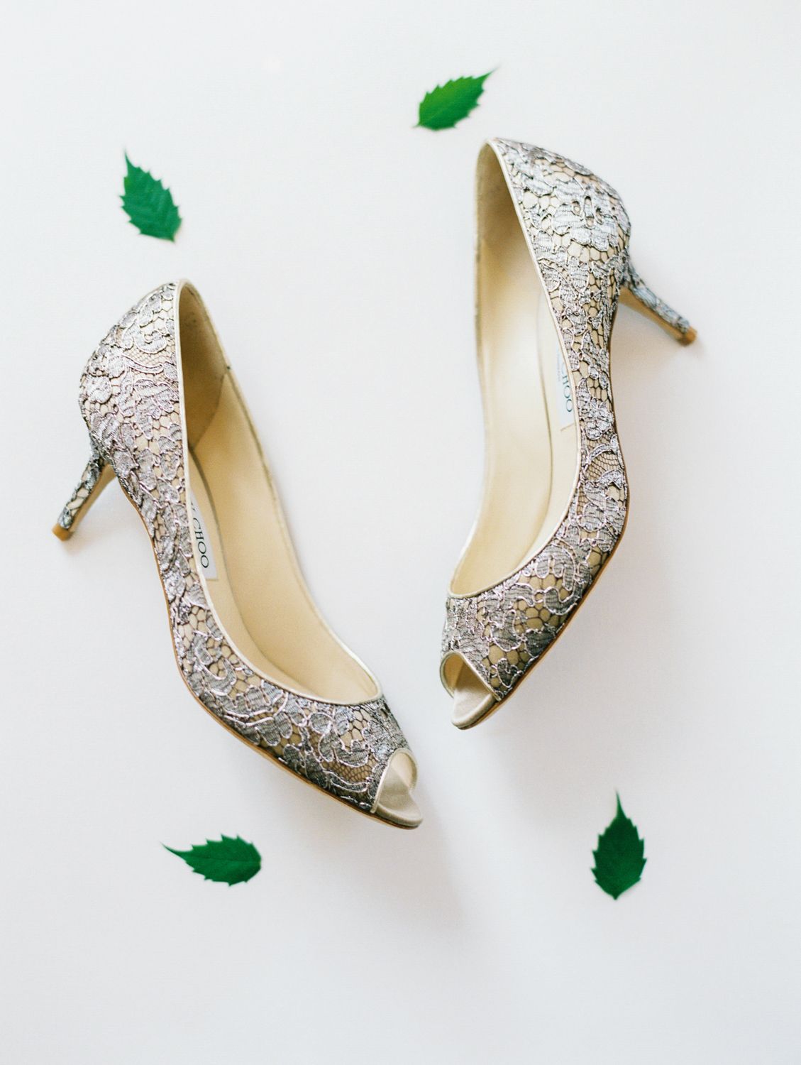 Gold lace peep-toe heels displayed on white background with scattered green leaves, elegant bridal wedding shoes.