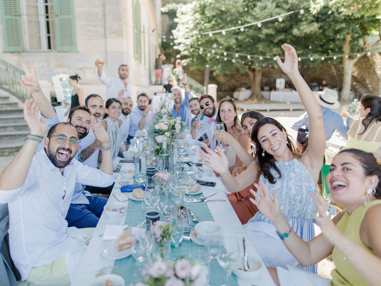 Joyful wedding guests celebrating at an outdoor long table dinner in a sunny Mediterranean courtyard.