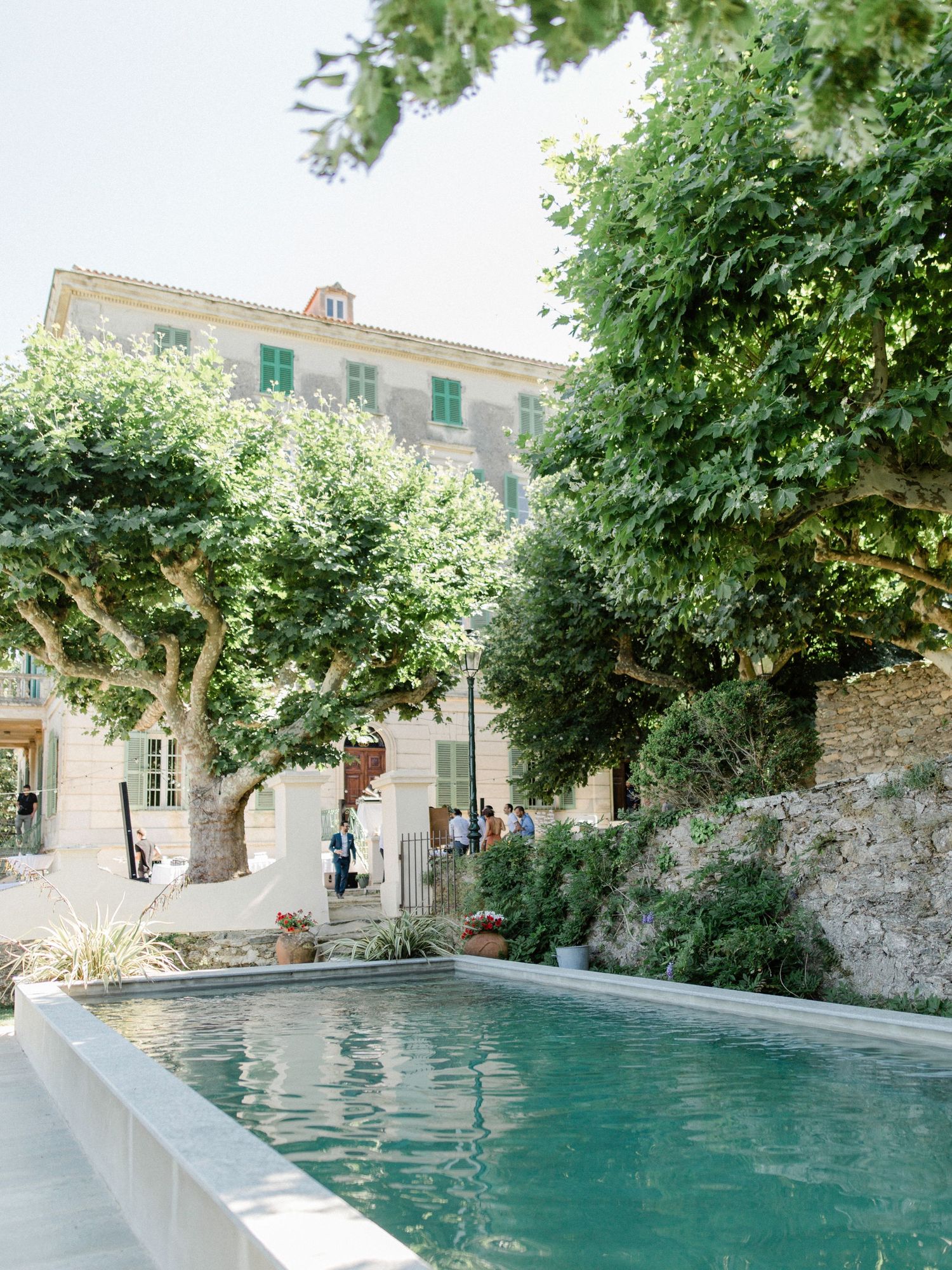 Elegant French villa with a serene pool, lush trees, and stone walls on a sunny day.