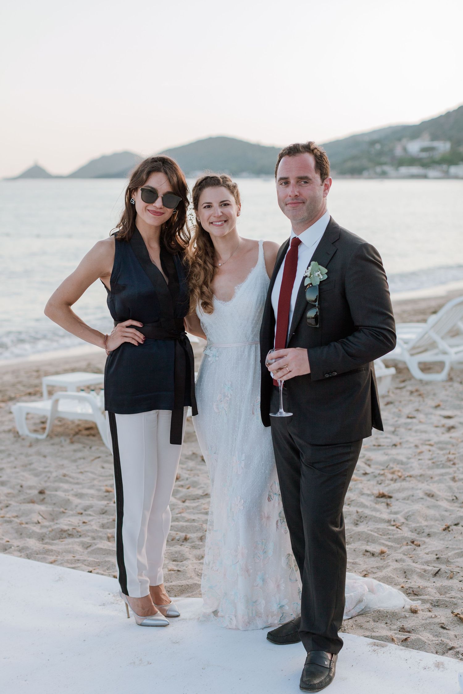 Les mariés posant avec une invitée sur une plage au coucher du soleil lors de la célébration de leur mariage.